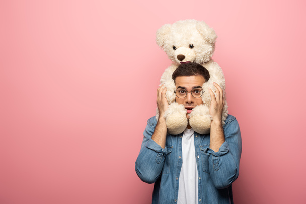 Scared young man holding teddy bear and looking at camera on pink background - Photo, Image