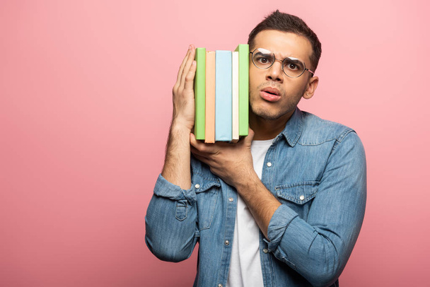 Thoughtful young man looking at camera while holding books on pink background - Foto, afbeelding
