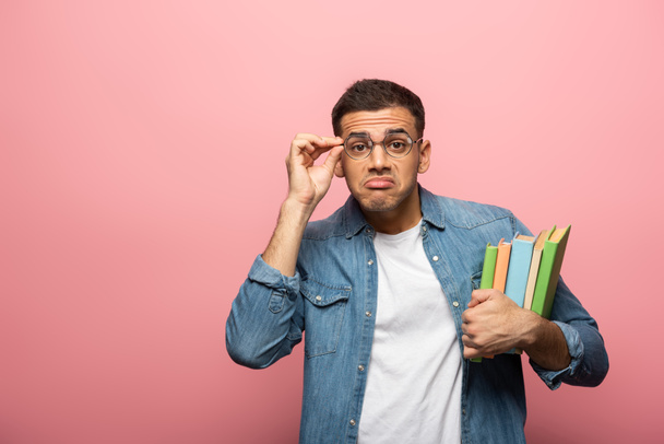 Young man grimacing while adjusting eyeglasses and holding books on pink background - Фото, зображення