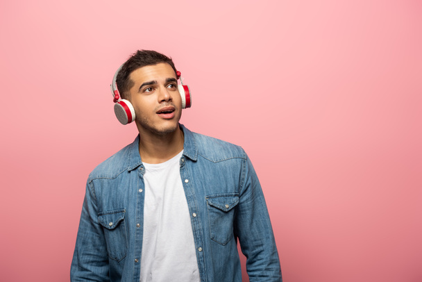 Dreamy young man in wireless headphones looking away isolated on pink - 写真・画像