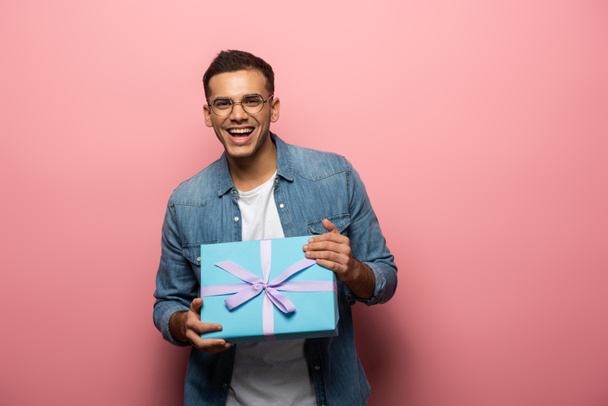 fröhlicher Mann lächelt in die Kamera und hält Geschenkbox auf rosa Hintergrund - Foto, Bild