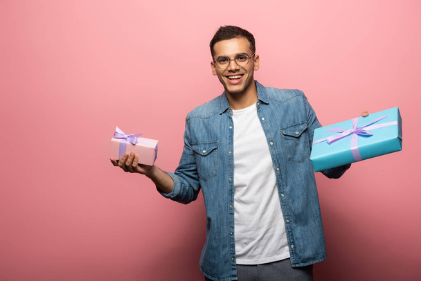 Smiling young man looking at camera while holding gift boxes on pink background - Valokuva, kuva