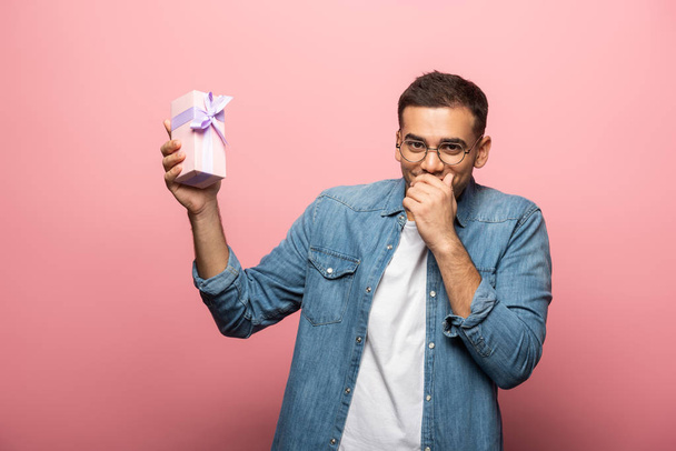 Positive man with hand by mouth holding gift box on pink background - Foto, immagini