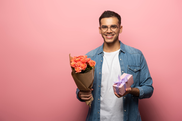 Young man with bouquet and gift smiling at camera on pink background - Photo, image