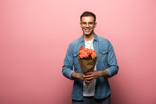 Handsome man smiling at camera while holding bouquet on pink background - Фото, зображення