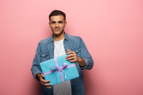 Young man looking at camera and holding gift with ribbon on pink background - Foto, Bild
