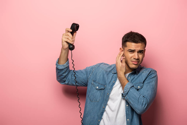 Hombre confuso con auricular telefónico que cierra la oreja sobre fondo rosa
 - Foto, Imagen