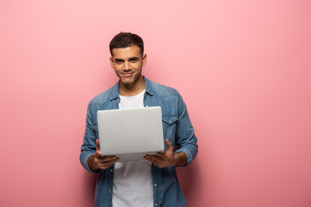 Handsome man holding laptop and smiling at camera on pink background - Φωτογραφία, εικόνα