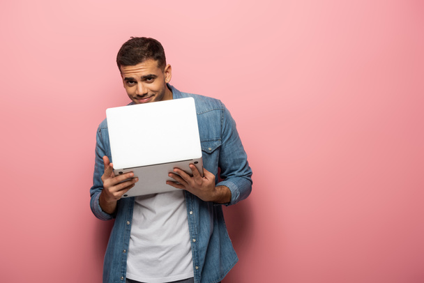 Fiatal férfi mosolyog a kamera, miközben tartja laptop rózsaszín háttér - Fotó, kép