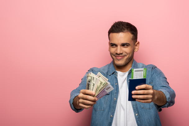 Улыбающийся мужчина показывает деньги и паспорт с посадочным талоном на розовом фоне
 - Фото, изображение