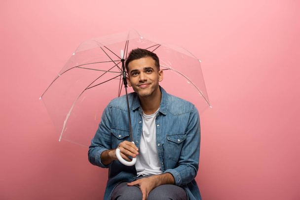 Smiling man holding transparent umbrella on pink background - Foto, imagen
