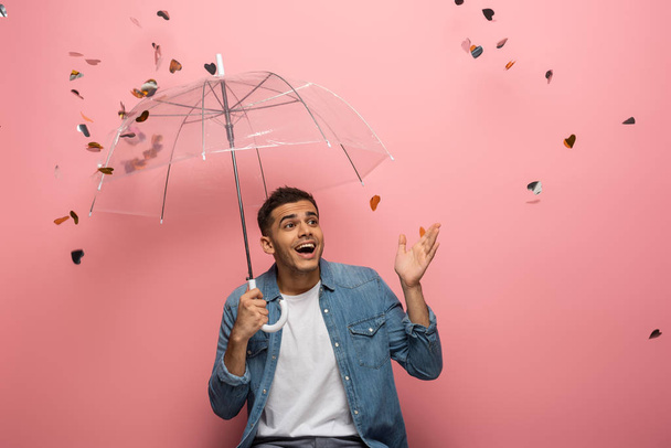 Surprised man with umbrella pointing with hand under falling confetti on pink background - Photo, Image