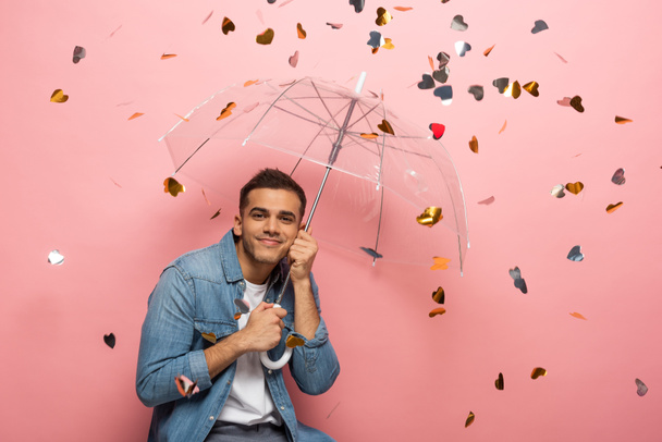 Young man with umbrella smiling at camera under falling heart shaped confetti on pink background - Foto, immagini