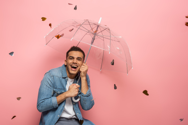 Allegro uomo che tiene l'ombrello sotto la caduta a forma di cuore coriandoli su sfondo rosa
  - Foto, immagini