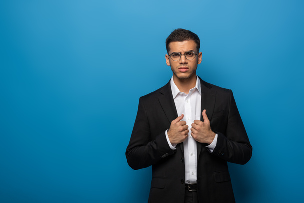 Confident businessman adjusting jacket and looking at camera on blue background - Foto, immagini