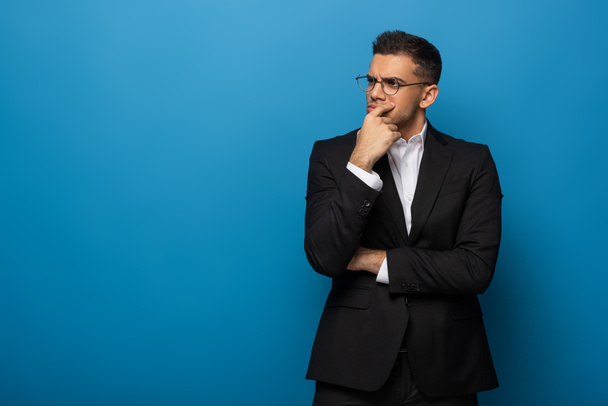 Thoughtful businessman with hand by chin looking away on blue background - Fotó, kép