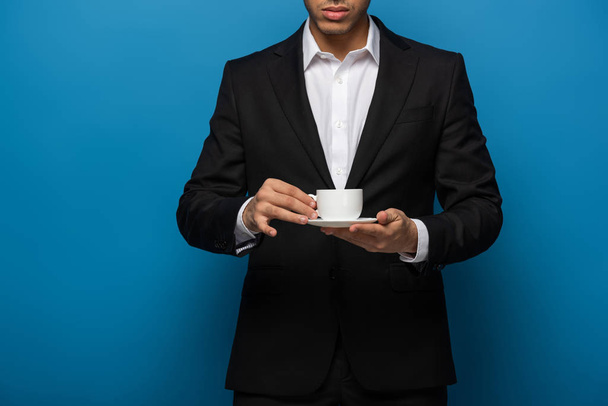 Cropped view of businessman holding coffee cup and saucer on blue background - Fotoğraf, Görsel