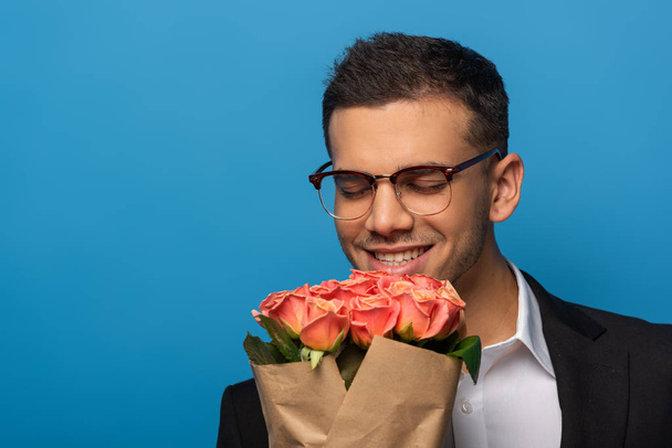 Smiling businessman with closed eyes holding bouquet isolated on blue - Foto, Bild
