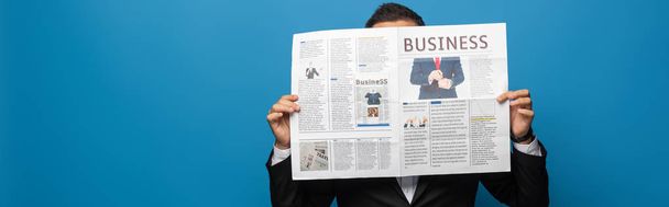 Панорамный снимок бизнесмена, закрывающего лицо во время чтения газеты, изолированной на голубом
 - Фото, изображение