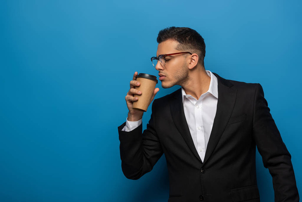 Vista lateral del joven empresario sosteniendo café para ir sobre fondo azul
 - Foto, imagen
