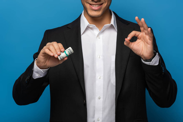 Vista cortada de homem de negócios sorridente mostrando gesto ok e segurando rolo de dinheiro no fundo azul
 - Foto, Imagem