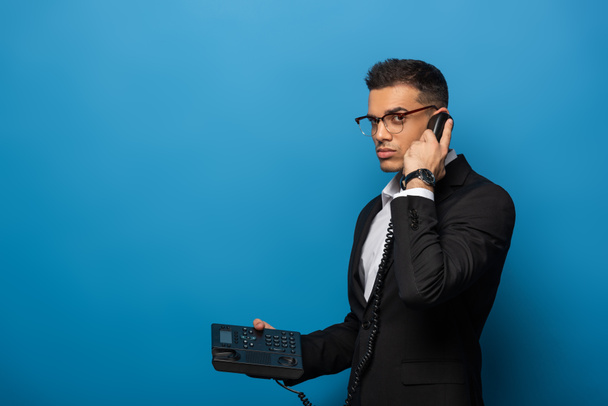 Вид збоку бізнесмена розмовляє по телефону і дивиться на камеру на синьому фоні
 - Фото, зображення