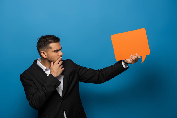 Side view of thoughtful businessman with hand by chin holding speech bubble on blue background - Foto, afbeelding