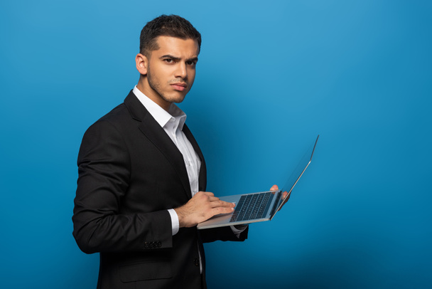 Side näkymä liikemies käyttää kannettavaa tietokonetta ja katsot kameraa sinisellä taustalla
 - Valokuva, kuva
