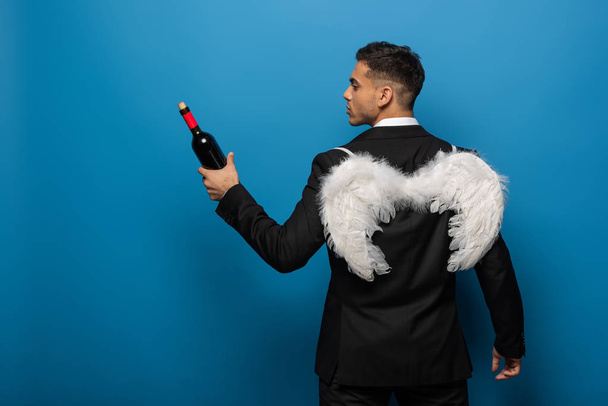 Back view of businessman with white wings holding bottle of wine on blue background - Valokuva, kuva