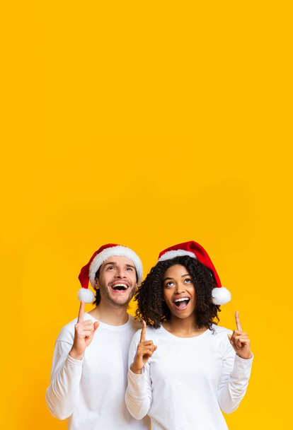 Vielrassiges Paar mit Weihnachtsmützen, die nach oben zeigen - Foto, Bild