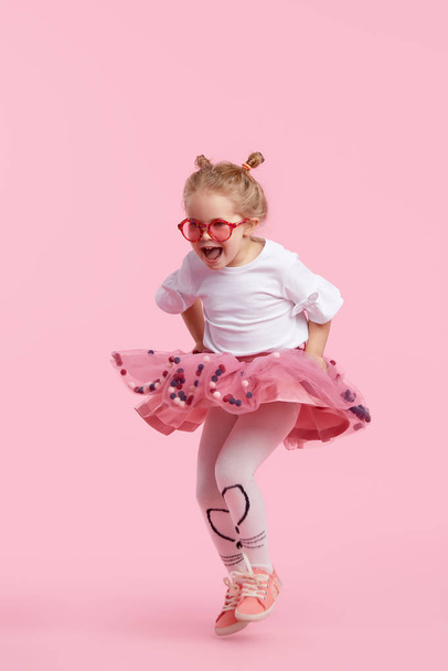 Happy childhood. Funny child girl in tulle skirt jumping and having fun isolated on pink background. Celebrating a vibrant carnival for kids, birthday - Foto, imagen