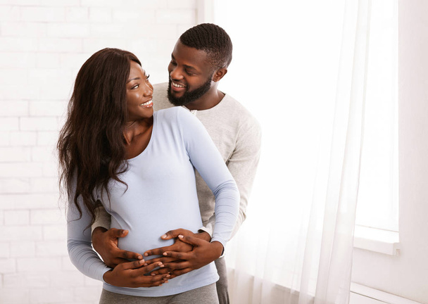 Coppia afro incinta che abbraccia accanto alla finestra a casa
 - Foto, immagini