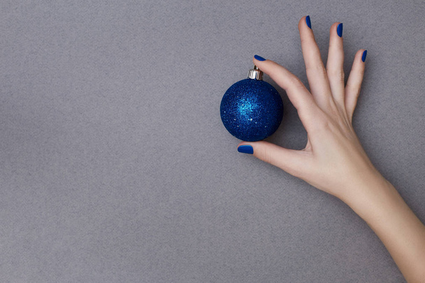 Female hand with manicure in trendy classic blue color holding sparkling blue christmas decorative ball on the grey background - Fotó, kép