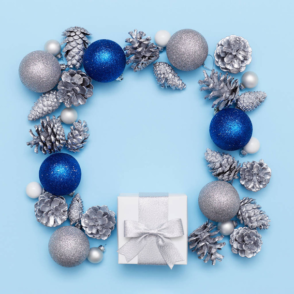 Flat lay frame composition of a white present box with silver box and silver fir cones and white, blue and silver balls on the blue background. Christmas concept - Zdjęcie, obraz