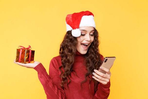 Χριστουγεννιάτικα ψώνια. Χαριτωμένη γυναίκα κρατώντας τηλέφωνο και δώρο στο καπέλο santas. - Φωτογραφία, εικόνα