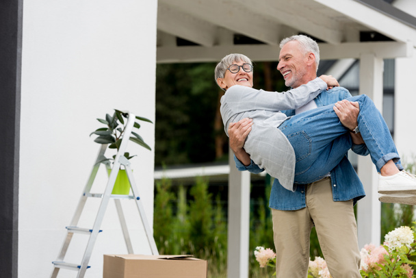 зрелый мужчина держит улыбающуюся женщину в очках рядом с новым домом
  - Фото, изображение