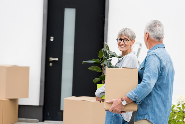 hombre maduro sosteniendo caja y mujer sonriente sosteniendo planta cerca de casa nueva
  - Foto, Imagen