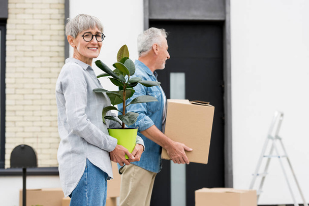 hombre maduro sosteniendo caja y mujer sosteniendo planta cerca de casa nueva
  - Foto, Imagen