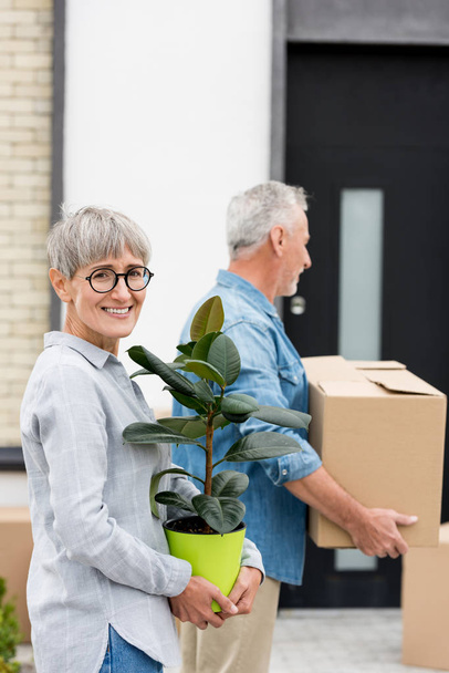 foco seletivo de mulher segurando planta perto e maduro homem segurando caixa perto de nova casa no fundo
  - Foto, Imagem