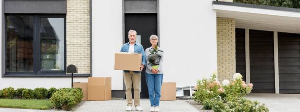 panoráma lövés érett férfi holding box és nő holding növény közelében új ház  - Fotó, kép