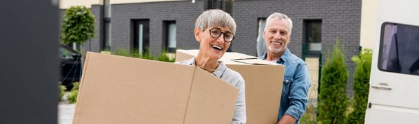 panoramiczne ujęcie dojrzałego mężczyzny i uśmiechniętej kobiety przynoszącej pudełka do nowego domu  - Zdjęcie, obraz
