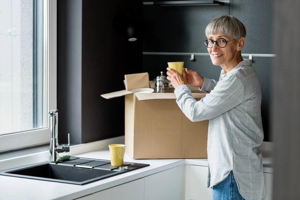 smiling mature woman unpacking box in new house  - Foto, afbeelding