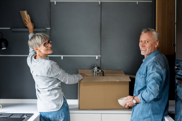 mature man and smiling woman unpacking box in new house  - Photo, Image