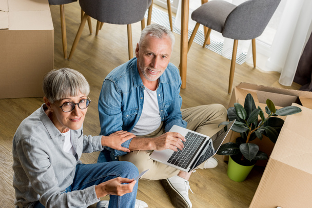 высокоугольный вид зрелого мужчины с ноутбуком и женщины, смотрящей на камеру в новом доме
  - Фото, изображение