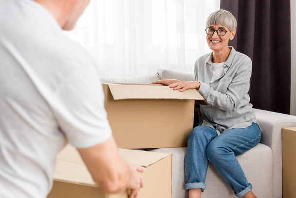 成熟した男が箱を持ち笑顔の女性が新しい家の中でソファに座って  - 写真・画像