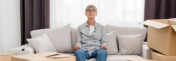 Panoramaaufnahme einer reifen Frau, die auf einem Sofa sitzt und in einem neuen Haus meditiert  - Foto, Bild