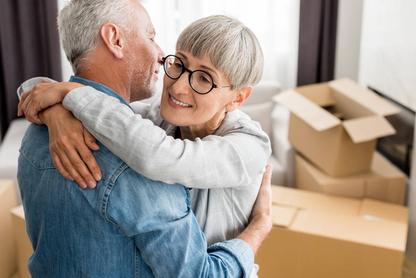 χαμογελαστή γυναίκα με γυαλιά αγκαλιάζει ώριμο άντρα στο νέο σπίτι  - Φωτογραφία, εικόνα