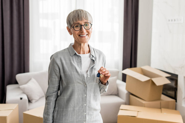 smiling woman holding keys and looking at camera in new house  - Photo, Image