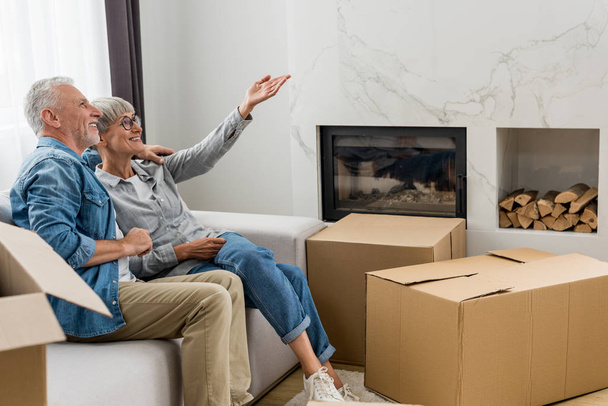 mature man and woman sitting on sofa and pointing with hand in new house  - Fotó, kép