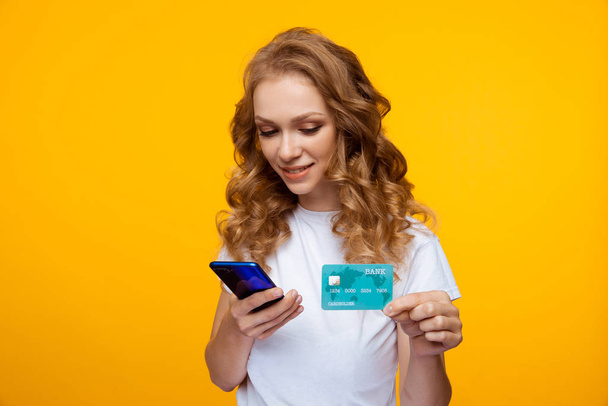 Conceito de compras online. Mulher bonita jovem segurando telefone e cartão de crédito azul em pé no estúdio amarelo
. - Foto, Imagem
